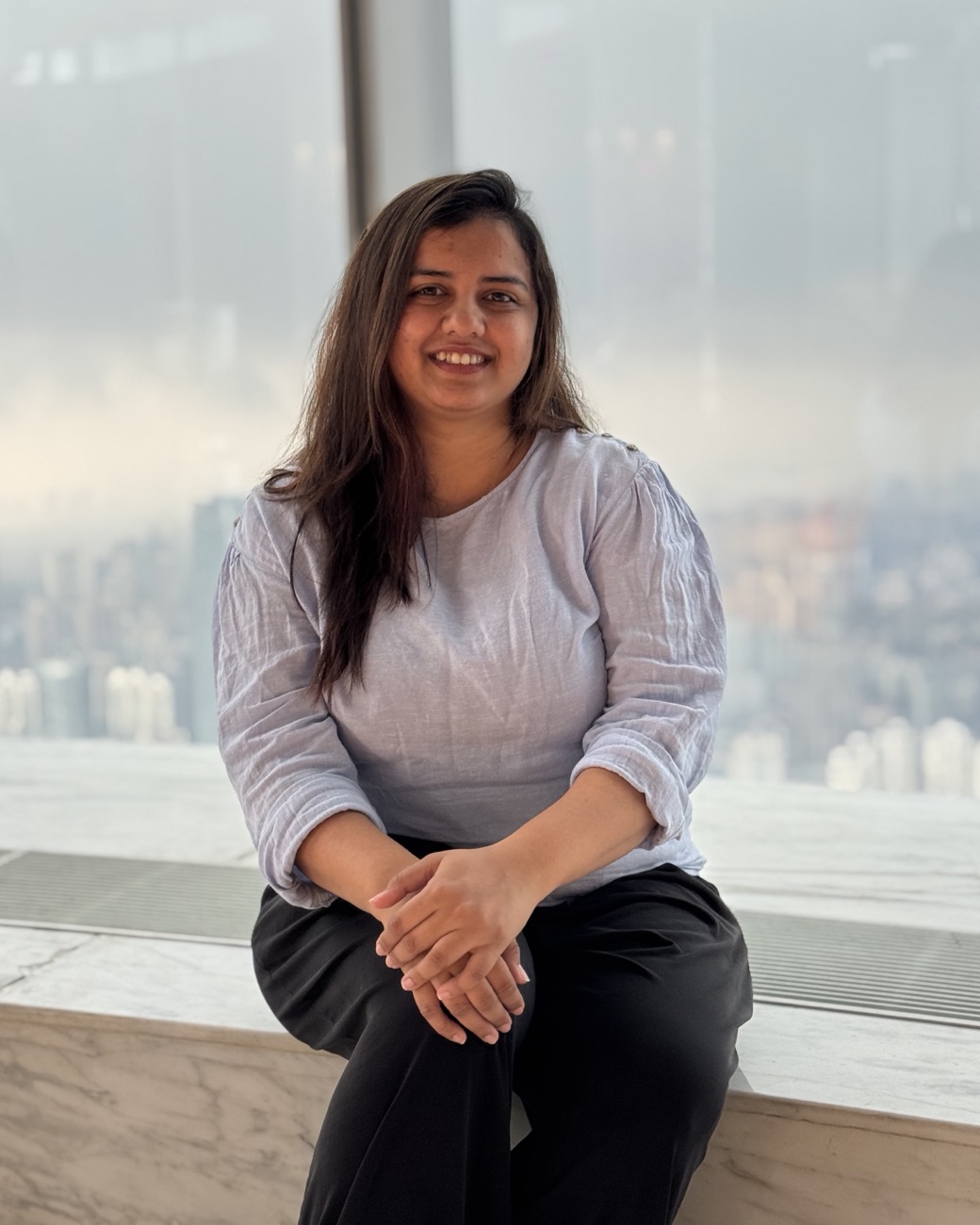 Aarti Kava, smiling, seated by a window high above a city.